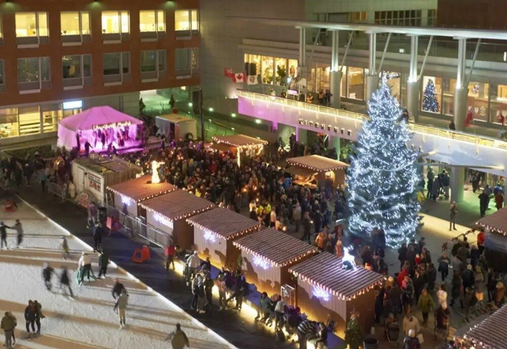 A festive outdoor scene featuring a large decorated Christmas tree surrounded by a bustling crowd. There are wooden market stalls offering holiday goods, with colorful lights illuminating the area at night. People skate on an ice rink nearby, enjoying the holiday atmosphere.