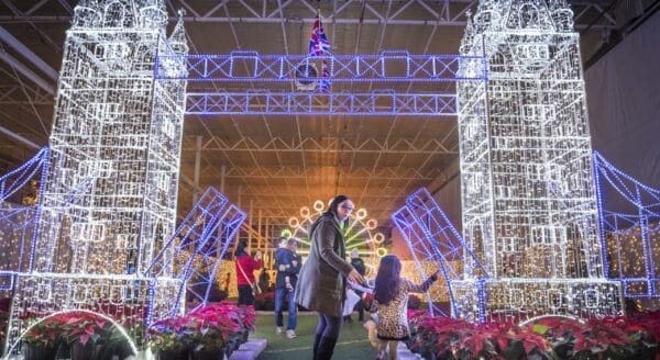 A woman and a child play under a dazzling, illuminated arch resembling a landmark, with vibrant flowers surrounding them. The scene is festive and magical, exuding a warm holiday atmosphere.