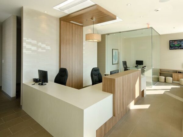 A modern reception area featuring a sleek front desk, two office chairs, and a welcoming seating space. Soft natural light illuminates the room, complemented by minimalist decor and a touch of greenery. The design emphasizes openness and professionalism.