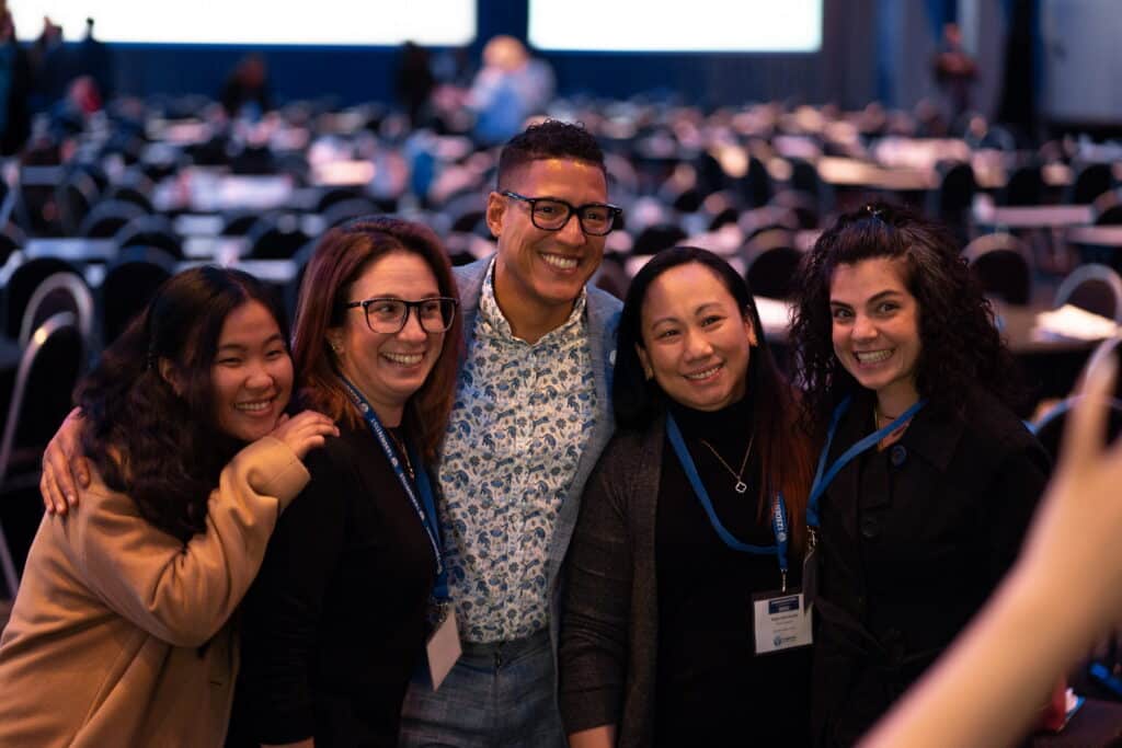 A group of five smiling individuals pose together for a photo at an event, with tables and people visible in the background. They appear friendly and engaged, showcasing a sense of camaraderie and enjoyment. The scene exudes positivity and connection.