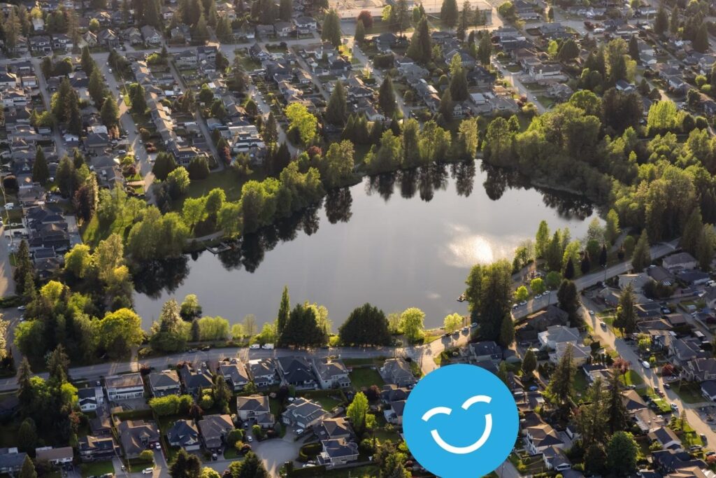 Aerial view of a suburban neighborhood with a central lake surrounded by houses and trees, under a sunlit sky.