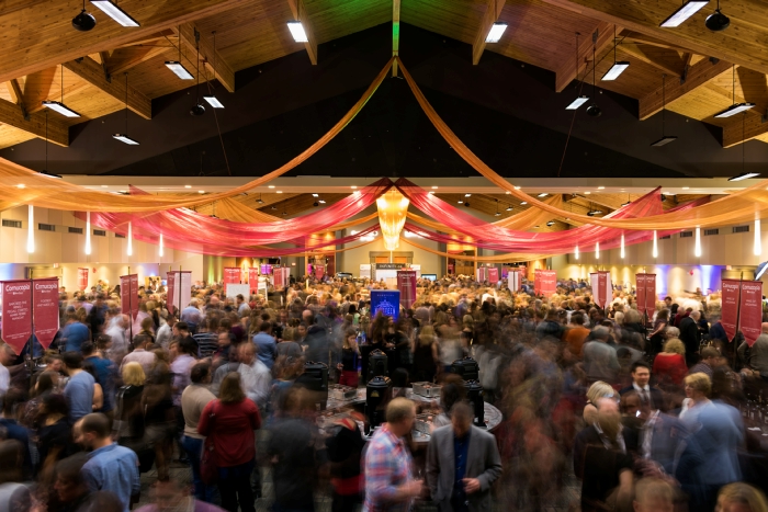 A lively indoor event featuring a large crowd of people gathered under colorful drapery. The scene shows vibrant decorations and an energetic atmosphere, indicating a social gathering or celebration.
