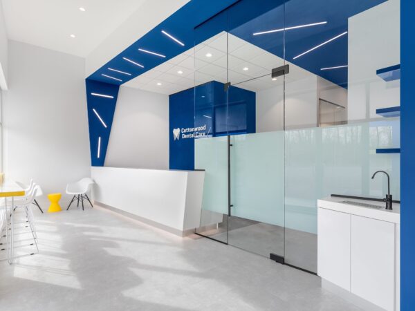 Modern office entrance with blue accents, glass door, and a white counter. Minimalist decor includes white chairs and a yellow bin.