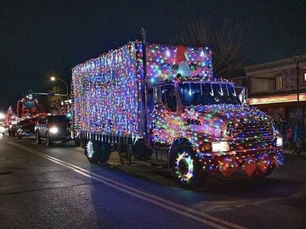 A brightly lit truck adorned with colorful holiday lights drives down a street at night, surrounded by festive decorations and other vehicles in a parade setting.