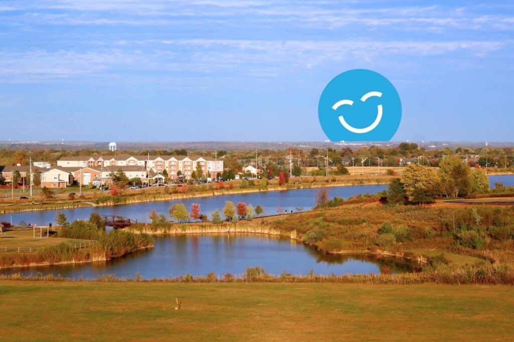 A peaceful landscape with calm water, autumn foliage, and a distant community. A blue smiley face symbol is prominent in the sky.