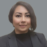 A professional woman with shoulder-length hair, wearing a dark blazer over a black top, smiles warmly at the camera. The background is a soft gray, enhancing her friendly demeanor.