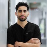 A young male medical professional in dark scrubs stands confidently with crossed arms. He is leaning against a gray wall in a well-lit healthcare environment.