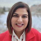 A woman with shoulder-length brown hair smiles warmly while wearing a red coat over a white blouse. The background features a snowy landscape, suggesting a winter setting.