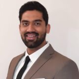 A smiling man wearing a light brown suit, white shirt, and black tie stands against a neutral background, exuding confidence and professionalism.