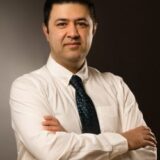 A man in a white dress shirt and patterned tie stands confidently with his arms crossed, posing against a dark, neutral background.