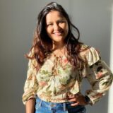 A young woman stands smiling, wearing a floral blouse and jeans. Sunlight highlights her hair and the textures of her outfit, creating a warm and inviting atmosphere.