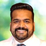 A man with a beard smiles warmly at the camera, wearing a white shirt and a blue tie. The background is softly blurred, featuring green and light colors, creating a cheerful ambiance.