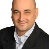 A smiling man with a bald head wearing a black blazer and a light blue striped shirt. He is positioned against a plain white background, conveying a professional appearance.