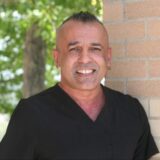 A smiling individual wearing dark scrubs stands next to a brick wall, with greenery in the background, suggesting a professional environment.