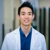 A young man wearing a white lab coat over a blue shirt smiles confidently in a well-lit indoor environment. He appears to be a medical professional.