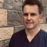 A man in scrubs stands in front of a stone wall, smiling confidently. He has short, dark hair and is looking at the camera.