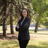 A woman standing confidently outdoors in a black blazer, with arms crossed, surrounded by trees and greenery. The background features a pathway and natural landscape, creating a vibrant and professional atmosphere.