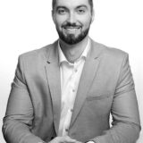 A smiling man with a beard is seated at a table, wearing a light gray suit and a white shirt. His hands are clasped in front of him, and he appears confident and approachable against a plain white background.