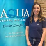 A smiling female dental professional in scrubs stands in front of a blue banner displaying "AQUA DENTAL WELLNESS" and the tagline "Dental Care for Life." The background features a textured stone wall and contact information for the dental clinic.