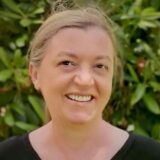A smiling woman with light brown hair, wearing a black top, is standing in front of lush green foliage. Her cheerful expression suggests a friendly demeanor.