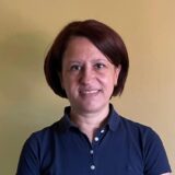 A woman with short brown hair is smiling and wearing a blue polo shirt, standing against a yellow background.