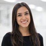 A young woman with long, dark hair smiles warmly at the camera. She is wearing a black shirt and stands against a softly blurred, bright background.
