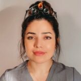 A woman with long dark hair styled in a bun stands against a neutral background, wearing a gray medical scrubs top. She has magnifying glasses resting on her head and a soft, neutral expression. The image conveys a professional appearance.