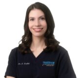 A smiling female dentist wearing a dark scrubs top with "Fraserview Dentist" embroidered on it. She has long, wavy hair and is standing with her arms crossed against a plain white background.