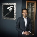 A professionally dressed individual stands confidently in front of a modern artwork against a dark wall. The person smiles, and their posture suggests approachability. The setting features a stylish entryway with a glass block wall.