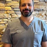 A smiling man wearing a gray medical scrub stands in front of a textured stone wall. He has a beard and short hair, exuding a professional demeanor.