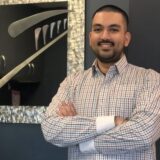 A man with short dark hair stands with his arms crossed, smiling in front of a stylized mirror with a modern design. He is wearing a checked shirt. The background features a dark wall that contrasts with the reflective mirror.
