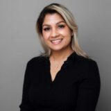 A smiling woman with medium-length blonde hair styled in soft waves, wearing a black shirt, poses against a gray background.