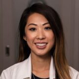 A smiling woman in a professional white coat stands against a neutral background. Her long, dark hair is partially wavy, and she has a warm, approachable expression. The image conveys a sense of friendliness and professionalism.