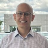 A smiling individual with a bald head and glasses stands in front of a modern building, wearing a checkered shirt. The background includes parked cars and a partly cloudy sky.