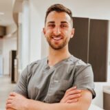 A smiling young man in scrubs stands with his arms crossed in a professional environment, likely a healthcare setting, conveying confidence and approachability.