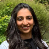 A woman with long dark hair smiles warmly at the camera, wearing a white coat over a black top. She stands outdoors surrounded by greenery, with a soft background that suggests a natural setting.