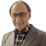 A smiling older man wearing glasses and a gray blazer over a checked shirt. He has a friendly expression and is posed against a plain white background.