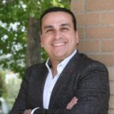 A smiling man stands with his arms crossed against a brick wall, greenery in the background. He wears a dark blazer over a white shirt, exuding a confident and approachable demeanor.