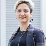 A woman with short, stylish hair smiles confidently while wearing a black top and a patterned jacket. She has a necklace and is accessorized with a camera strap around her neck. The background features soft, blurred lines.