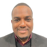 A smiling man wearing a black suit and a maroon shirt stands against a white background, exuding professionalism and friendliness.