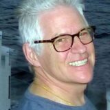 A smiling man with salt-and-pepper hair and glasses stands against a backdrop of water, exuding a friendly and approachable demeanor. He wears a casual gray shirt and appears to be enjoying a relaxed moment.