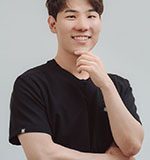 A young man with short, dark hair smiles while resting his chin on his hand. He wears a black shirt and stands with his arms crossed against a light gray background.