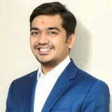 A smiling man wearing a blue blazer and white shirt stands against a neutral background. He has a small decorative mark on his forehead.