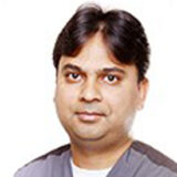 A smiling male dentist with dark hair and wearing dental scrubs is looking at the camera against a white background.