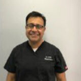 A smiling dental professional wearing a black coat is standing against a neutral background. He appears friendly and approachable, likely conveying a welcoming atmosphere in a dental setting.