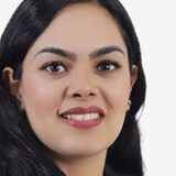 A close-up portrait of a smiling woman with long dark hair and a professional appearance, wearing a black blazer. She has striking eyes and is set against a plain white background.