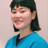 A smiling young woman with short black hair, wearing a teal medical scrub, poses for the camera in a professional setting.