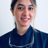 A smiling woman in a dark blue medical uniform stands in front of a light background. She wears dental loupes around her neck, indicating her profession in dentistry or healthcare.