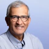 A smiling middle-aged man with short gray hair and glasses, wearing a light blue button-up shirt, poses against a soft blue background.