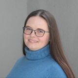 A young woman with long hair, wearing glasses and a blue turtleneck sweater, smiles gently while standing against a neutral-colored background.